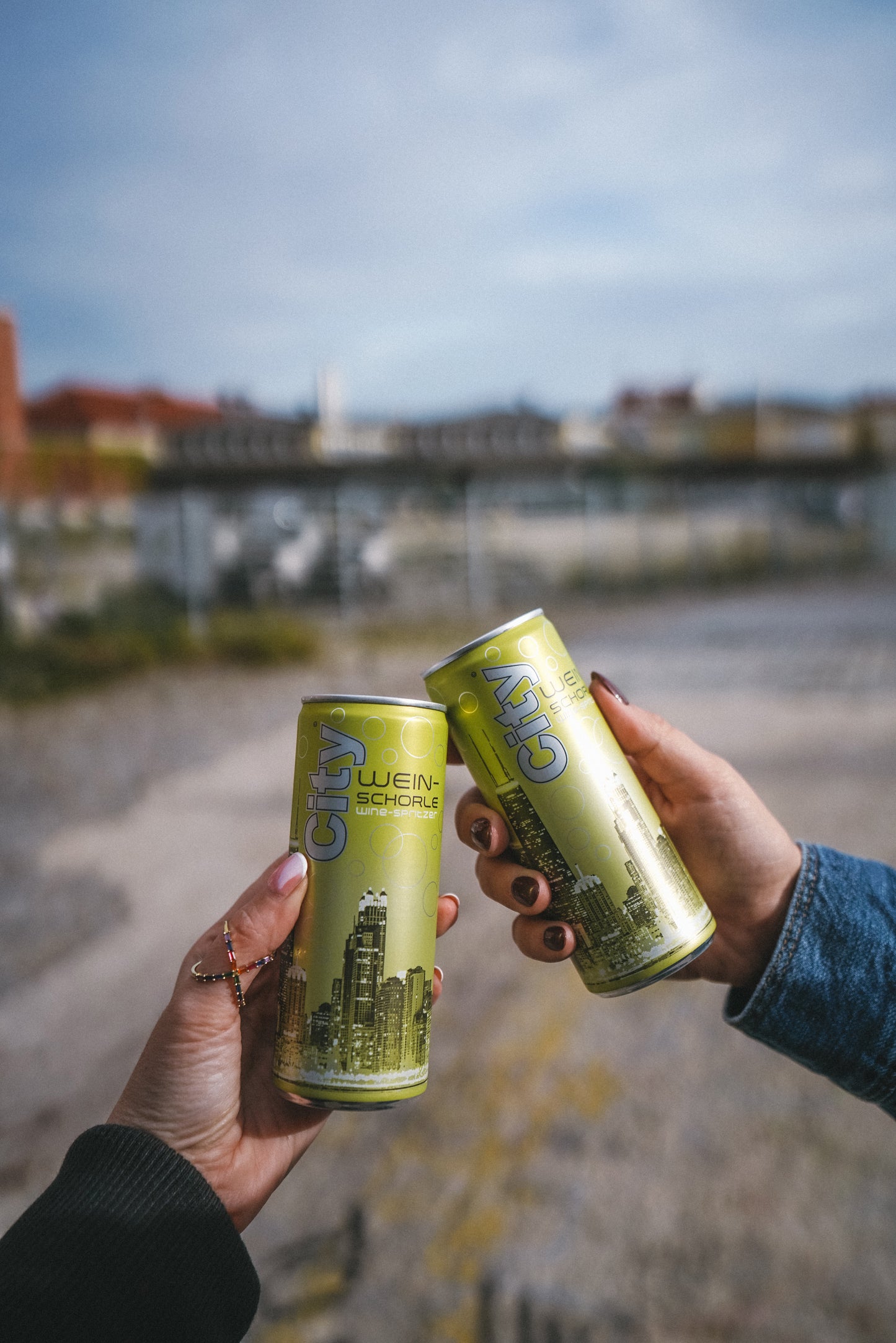 Zwei Dosen City Weinschorle  weiss gehalten von zwei Frauen Händen, Hintergrund urban aber verschwommen. Dose ist grün mit City Skyline 