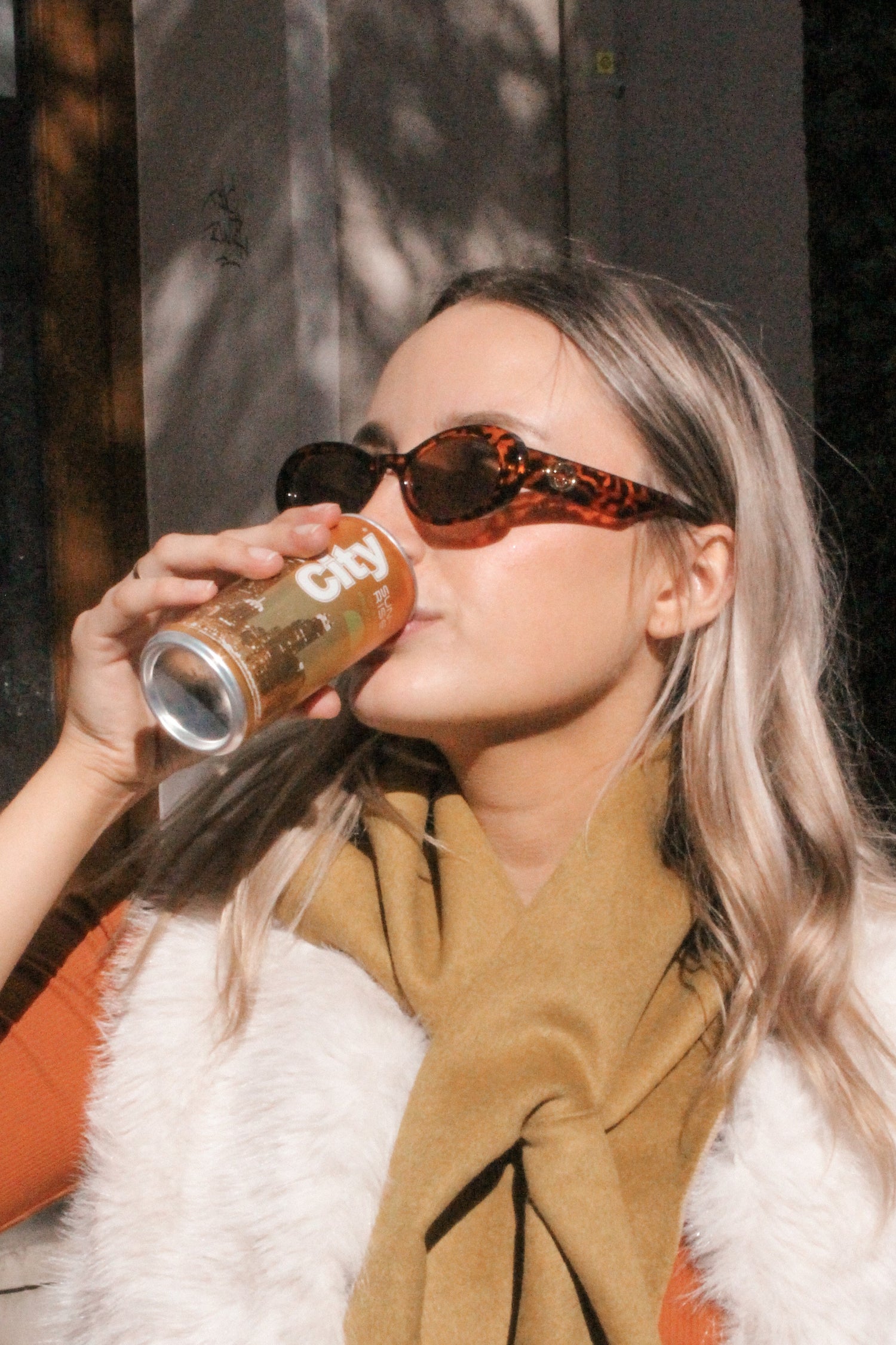 blonde Frau trink aus einer orangenen Dosen. Sie trägt einen Sonnenbrille und hat einen schal und ein Mantel an